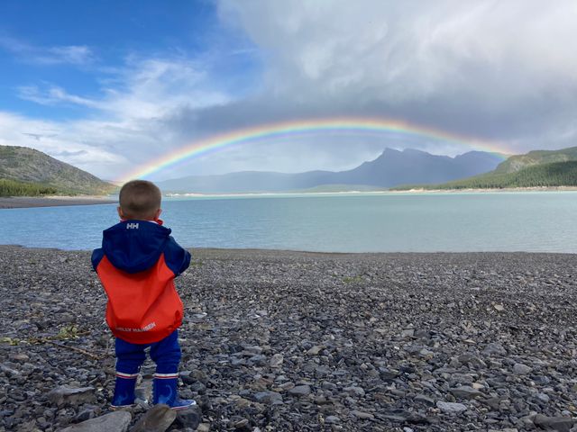Een peuter in een regenjas staat aan de oever van een bergmeer en kijkt naar een regenboog.